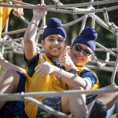  Two young boys in turbans giving thumbs up while climbing on a rope structure at a playground.