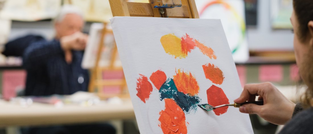 A student paints green onto a canvas filled with orange and yellow circles. The canvas is propped on an easel. Another student works in the distance.