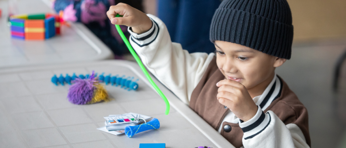 A young child in a beanie cap engaging with sensory play materials at a table during an indoor event.