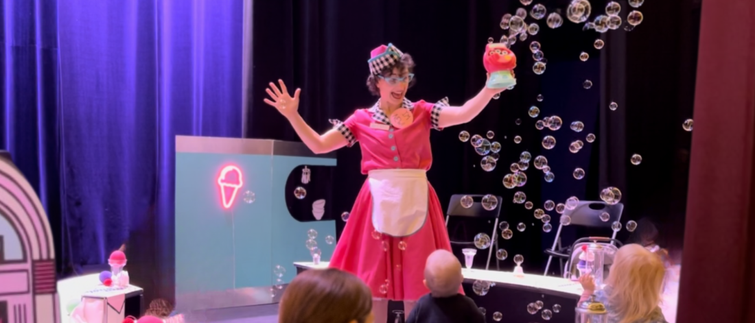 Actor dressed in a 1950s diner style waitress costume stands on the stage in front of audience while she holds a bubble making machine with bubbles coming out of it.